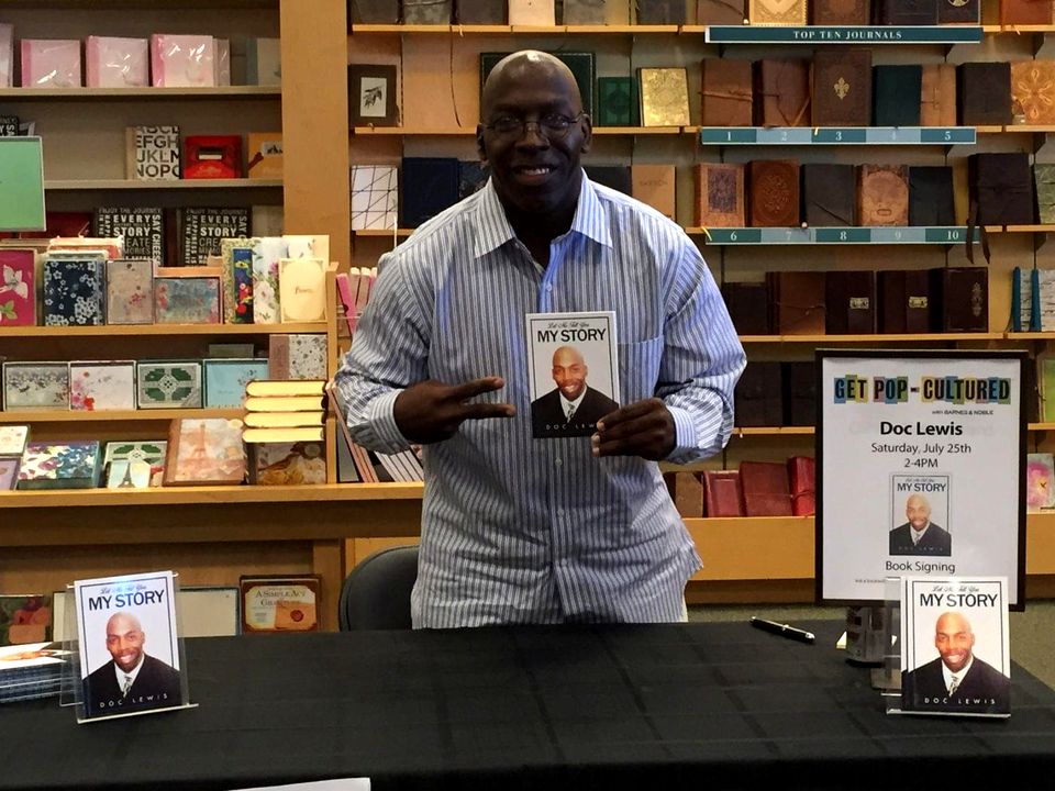 Doc Lewis at a bookstore signing table, holding up a copy of his book, Let Me Tell You My Story
