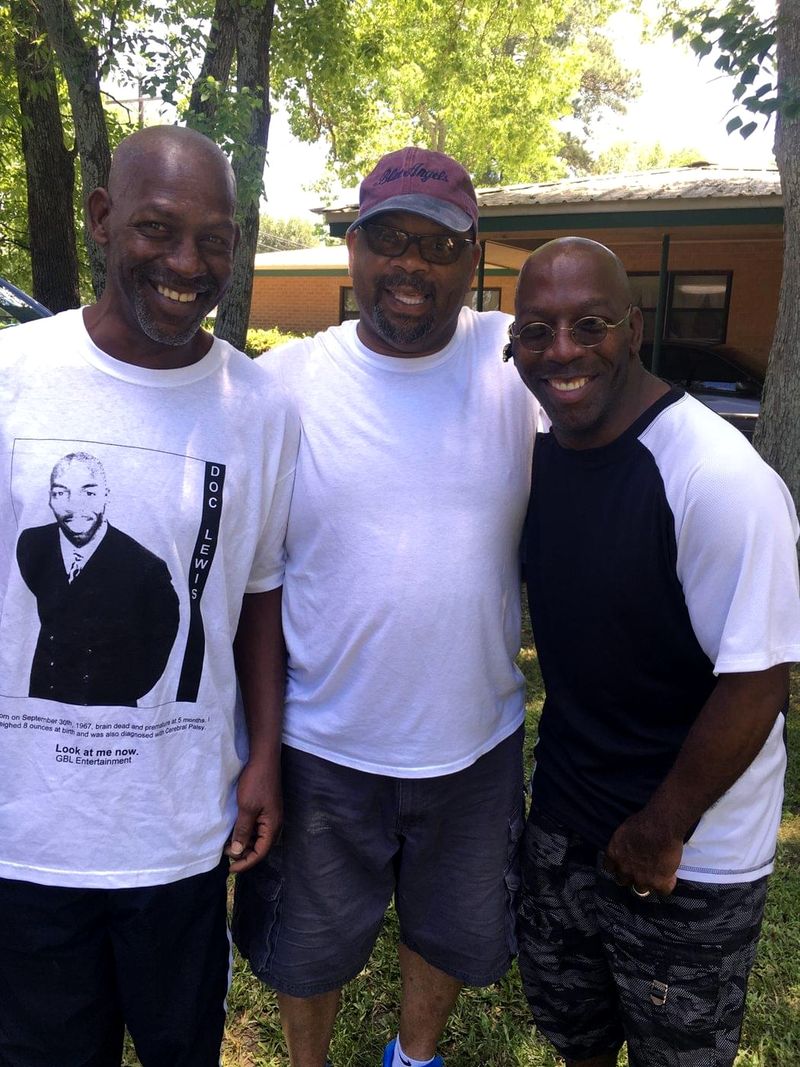 Doc Lewis with two friends outdoors, one wearing a T-shirt printed with Doc's photo and name