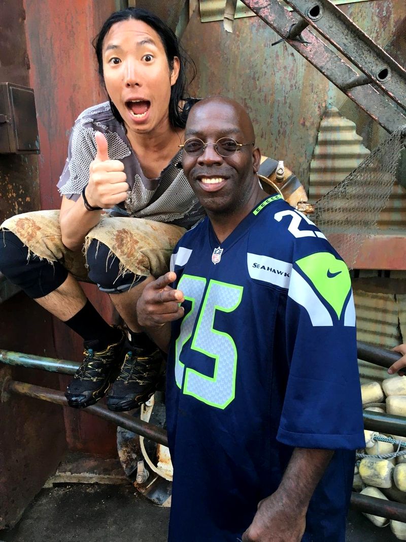 Doc Lewis laughing in a football jersey while a friend gives a thumbs up behind him