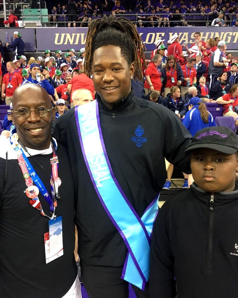 Doc Lewis with a smiling athlete wearing a sash and a boy in a cap, in a stadium full of athletes