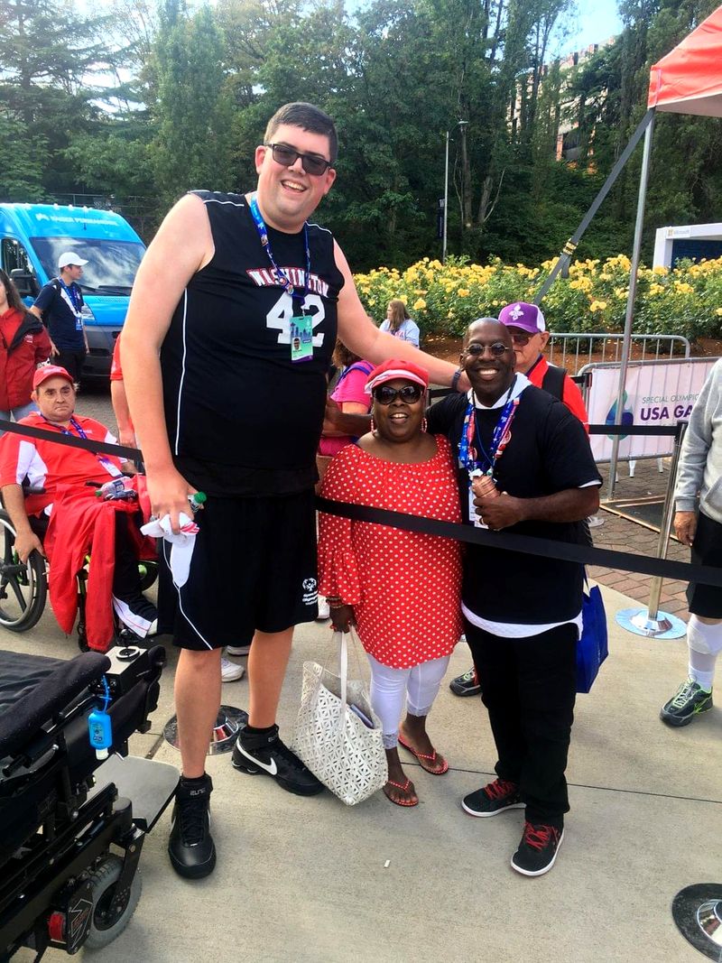Doc Lewis with a tall athlete and a woman in a red polka-dot dress beside a barrier